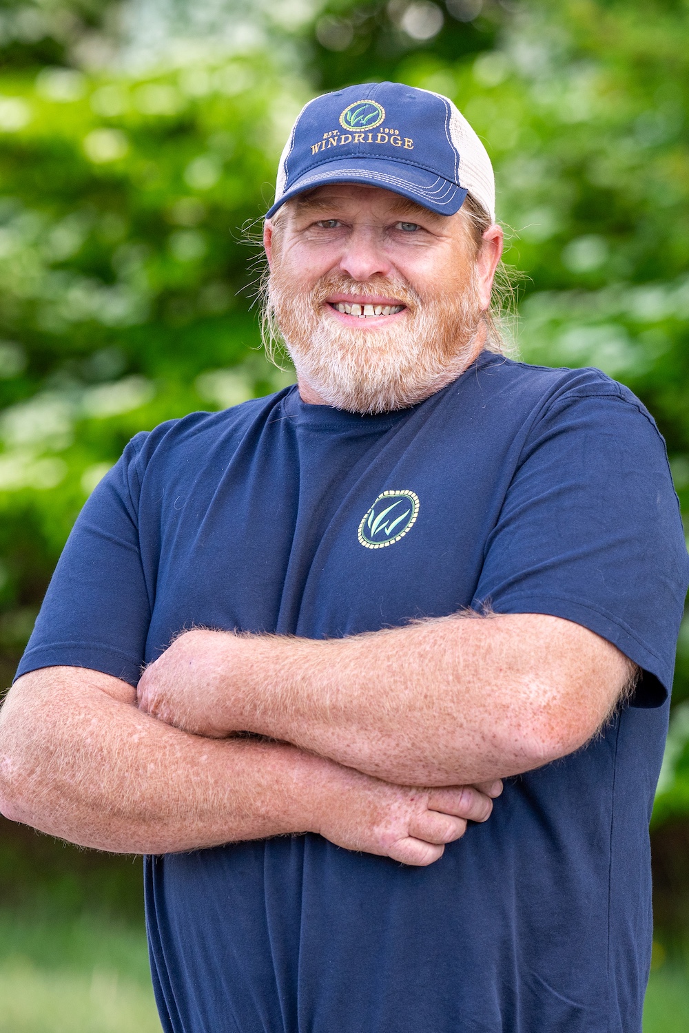 Man in blue Windridge shirt and cap smiling with folded arms outdoors in front of lush green background.