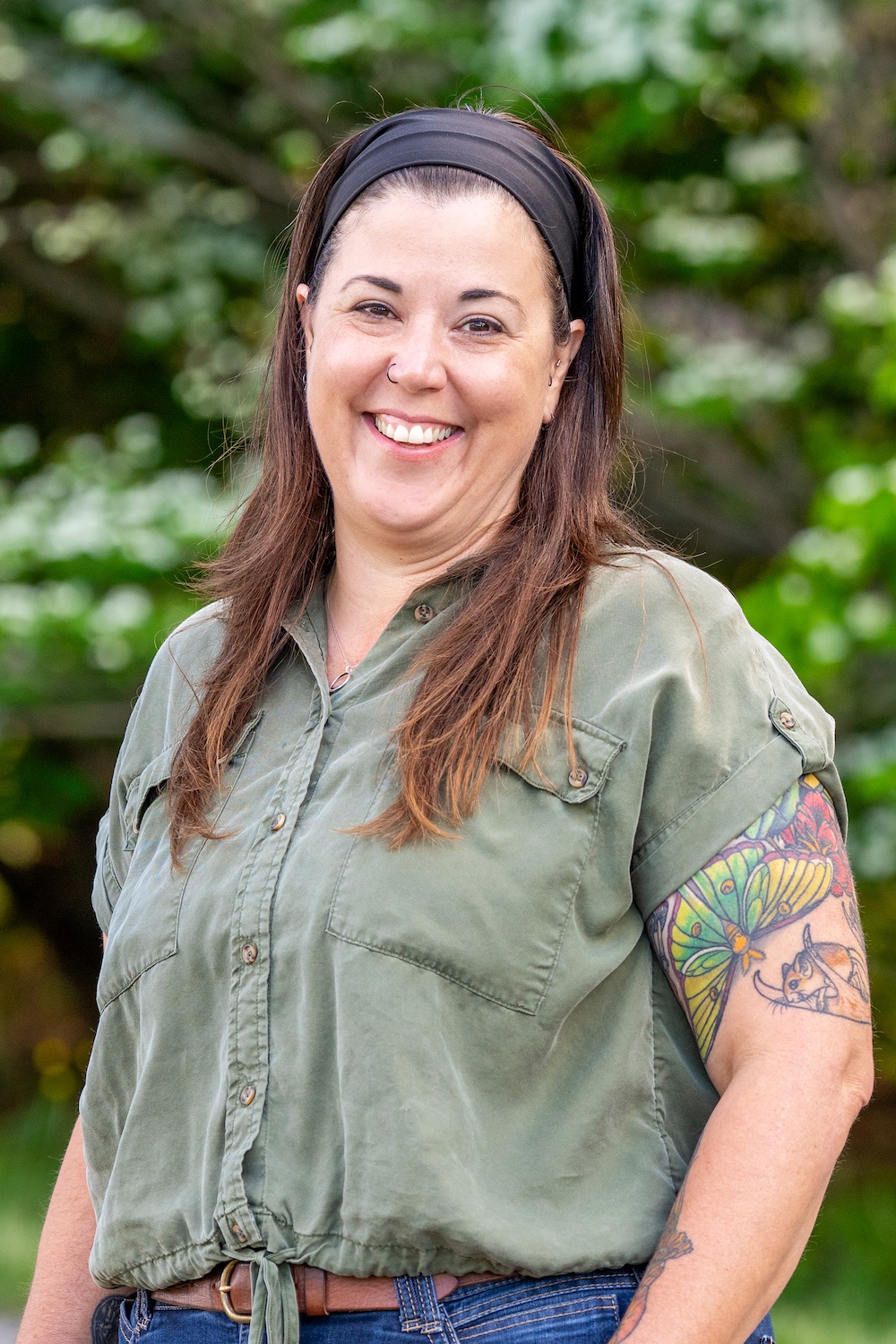 Smiling person with headband and tattoo in green shirt, standing outdoors against a blurred greenery background.