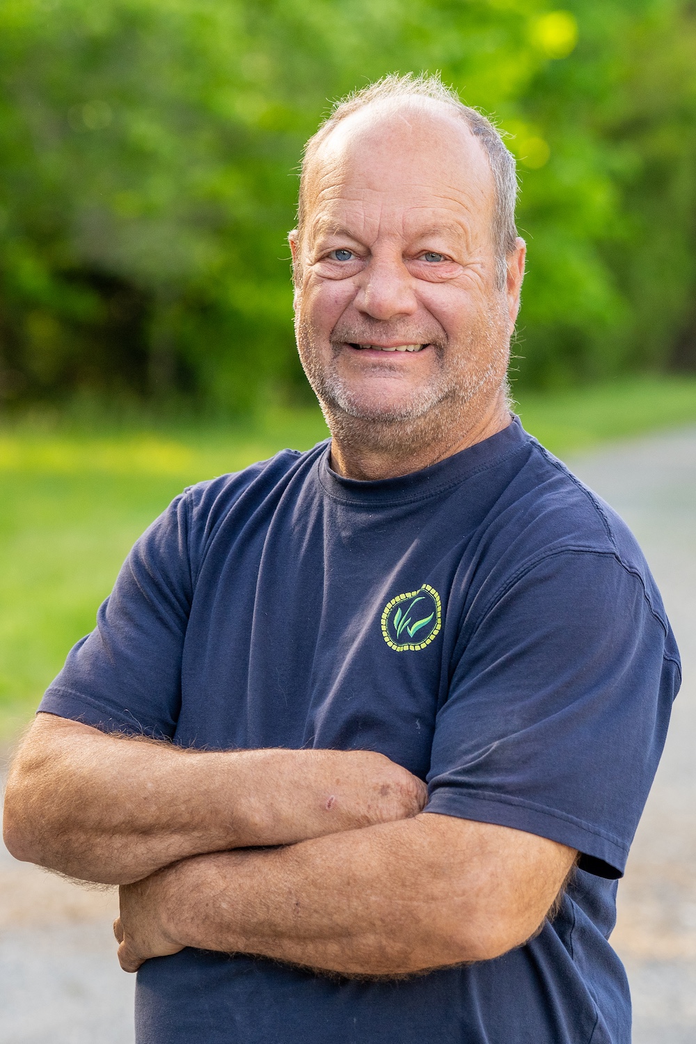 Smiling man in a navy shirt with folded arms standing outdoors with a green background.