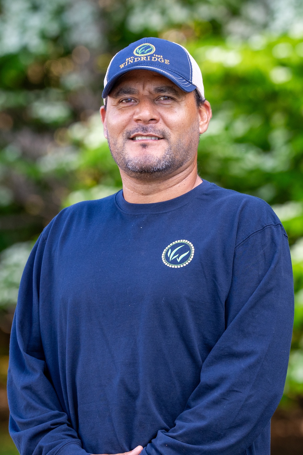 Smiling man in a navy cap and shirt with a logo, outdoors with blurred green background.
