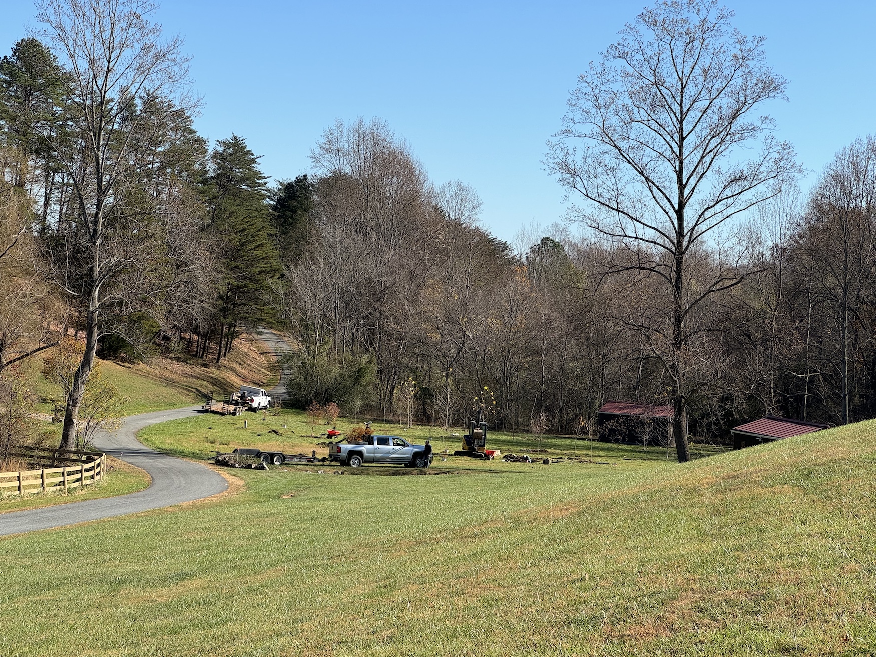 Winding country road with pickup trucks, bare trees, and grassy fields under a clear blue sky.